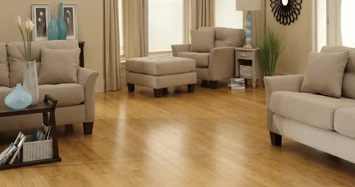 A living room with professionally installed bamboo flooring, beige sofas, a matching ottoman, a side table with a blue lamp, vases, framed pictures, and large windows with blinds and curtains in Memphis, TN